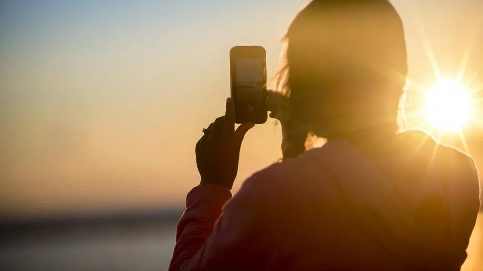Gün batımında arkadan görünen bir kadın elindeki telefonla manzarayı çekiyor