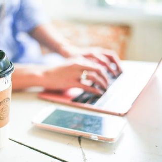 Kahve bardağının yanında dizüstü bilgisayarda yazı yazan eller ve bir telefon